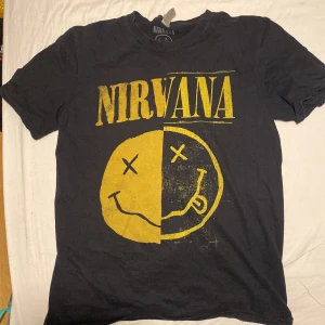 Svart Nirvana t-shirt från Gildan - Cool svart t-shirt från Gildan med det ikoniska Nirvana-trycket i gult på Framsidan. Perfekt för alla Nirvana-fans som vill visa sin kärlek till bandet !