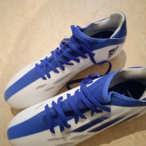 Blå och vita fotbollsskor från Adidas - Säljer ett par blå och vita fotbollsskor från Adidas. Skorna har en strömlinjeformad design med blå detaljer och Adidas-loggan på sidan. Perfekta för fotbollsplanen med sina dobbar för bra grepp.
