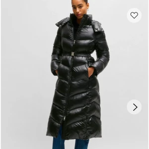 Svart lång dunjacka med bälte - Snygg och varm svart lång dunjacka med bälte i midjan. Jackan har en hög krage och huva för extra skydd mot kylan. Den är quiltad och har en dragkedja framtill. Perfekt för kalla vinterdagar.