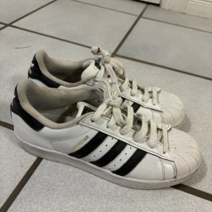 Vita Adidas Superstar sneakers - Klassiska vita Adidas Superstar sneakers med svarta ränder och snörning. Skorna har den ikoniska snäckformade tån och Adidas-loggan på hälen.