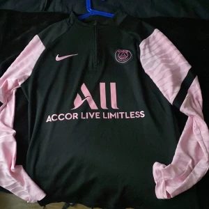 Svart och rosa fotbollströja från psg/nike - Snygg svart fotbollströja med rosa detaljer från Nike. Tröjan har långärmade rosa ärmar och  Perfekt för fotbollsfans som vill sticka ut!