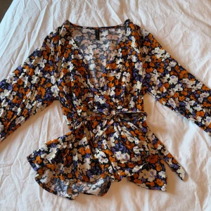 Blommig omlottblus med knytband - Säljer en färgglad blommig omlottblus med långa ärmar och knytband i midjan. Blusen har ett livligt mönster i orange, lila och vit. Perfekt för att liva upp garderoben!