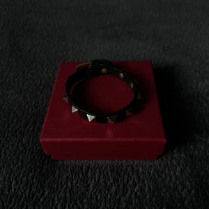 Svart armband med nitar från Valentino - Snyggt svart armband från Valentino med silverfärgade nitar. Kommer i originalförpackning med dustbag och extra nitar. Enda nackdelen är att armbandets ena ände har gått sönder (se bild), men går fortfarande att använda hur bra som helst och syns inte vid användning. Dessutom går det att limma fast änden. Kom privat vid funderingar