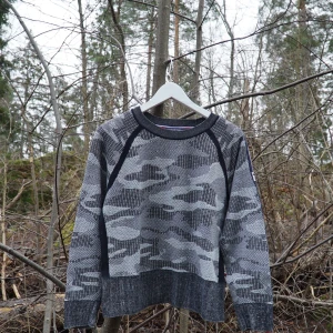 Grå camouflagemönstrad tröja från Tommy Hilfiger - 🌟Säljer en cool grå tröja från Tommy Hilfiger med camouflagemönster. Tröjan har långa ärmar och en ribbad krage🌟Detaljer i form av ränder i rött och vitt på ärmen ger en sportig touch. Perfekt för en avslappnad stil!🤌🏻❌Finns tho små fläckar, en på framsidan och en på ena armen❌ Går säkert lätt bort med tvätt✌🏻