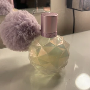 Parfym med lila fluffboll - Säljer en parfym från Ariana Grande med en unik flaska i geometrisk design och en lila fluffboll som dekoration. Flaskan har en ljusrosa kork och ger en lyxig känsla. Perfekt för den som älskar söta och fruktiga dofter.
