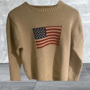 Beige stickad tröja från GANT - Säljer en beige stickad tröja från GANT med ett amerikanskt flaggmönster på framsidan. Tröjan har långa ärmar och ribbstickade detaljer vid hals, ärmslut och nederkant. Perfekt för en avslappnad stil.