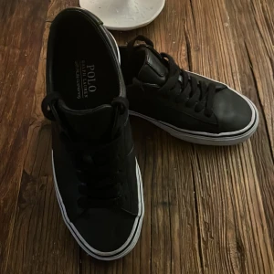 Svarta sneakers från Polo Ralph Lauren - Snygga svarta sneakers från Polo Ralph Lauren med klassisk design. Skorna har en vit sula och svarta snören, vilket ger en stilren look. Perfekta för en avslappnad stil.