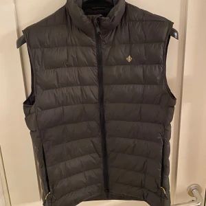 Olivgrön dunväst från Morris - Snygg dunväst från Morris med dragkedja och en liten broderad detalj på bröstet. Perfekt för kyligare dagar och ger en stilren look. Västen har en hög krage och är quiltad för extra värme. Jag säljer den för att det inte kom till användning. Köpte den i somras för 2300kr använd väldigt lite. Tveka inte på att höra av er om ni har några frågor! Fler bilder kan skickas om de önskas. Priset är inte hugget i sten.
