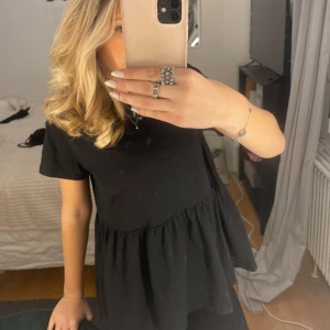 Svart t shirt med volangdetalj - Säljer en stilren svart blus med volangdetalj i nederkant. Blusen har korta ärmar och en lös passform, perfekt för en avslappnad look. Passar bra till både jeans och kjol.