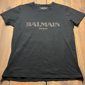 Svart t-shirt från Balmain - Snygg svart t-shirt från Balmain med tryck 'BALMAIN PARIS' på framsidan. Perfekt för en stilren look. T-shirten är kortärmad och har en klassisk passform. Stolen M är 185 och passar perfekt. 