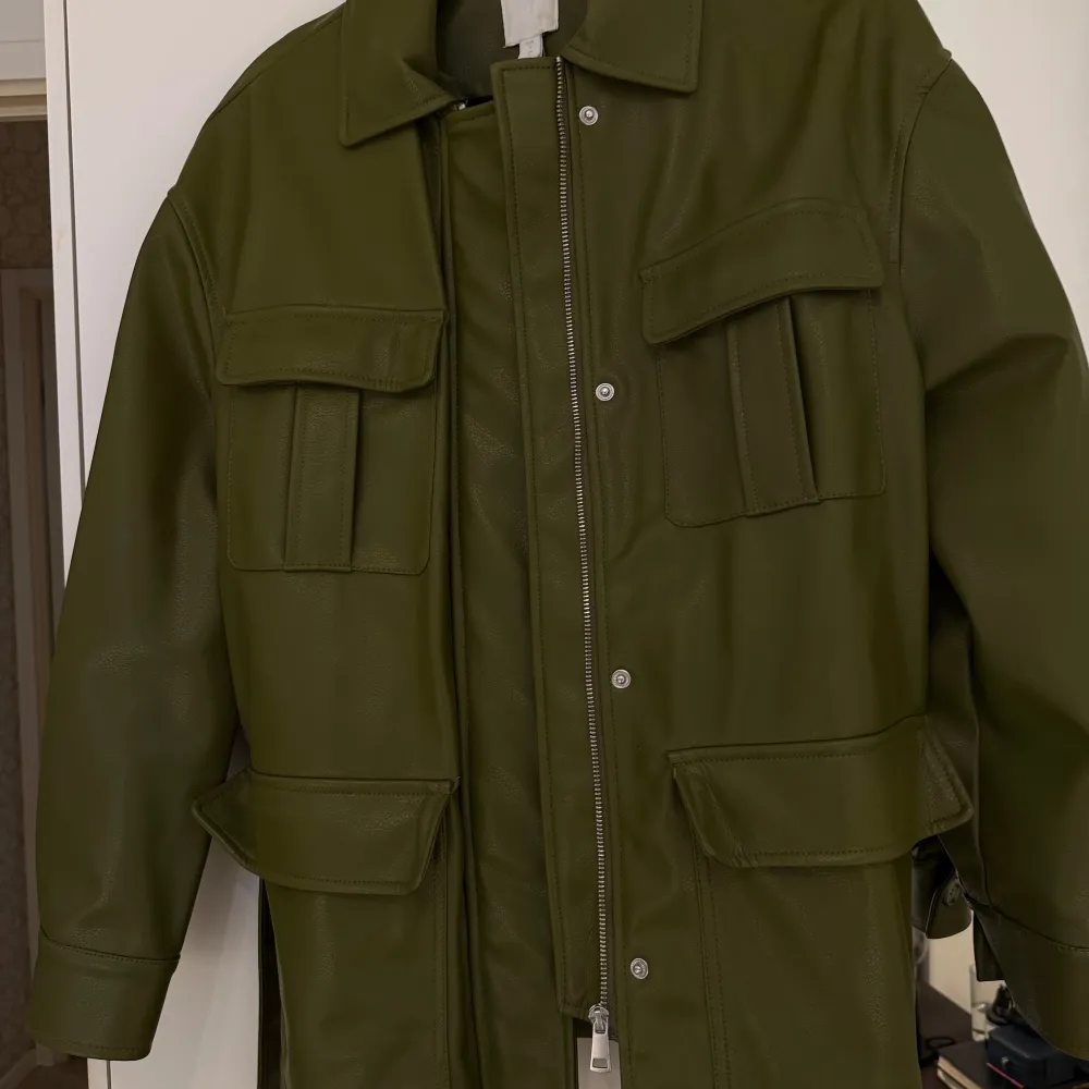 Snygg grön skinnjacka med knytbälte i midjan och knappar framtill. Jackan har långa ärmar och flera praktiska fickor. Perfekt för att ge en cool touch till din outfit.. Takit.