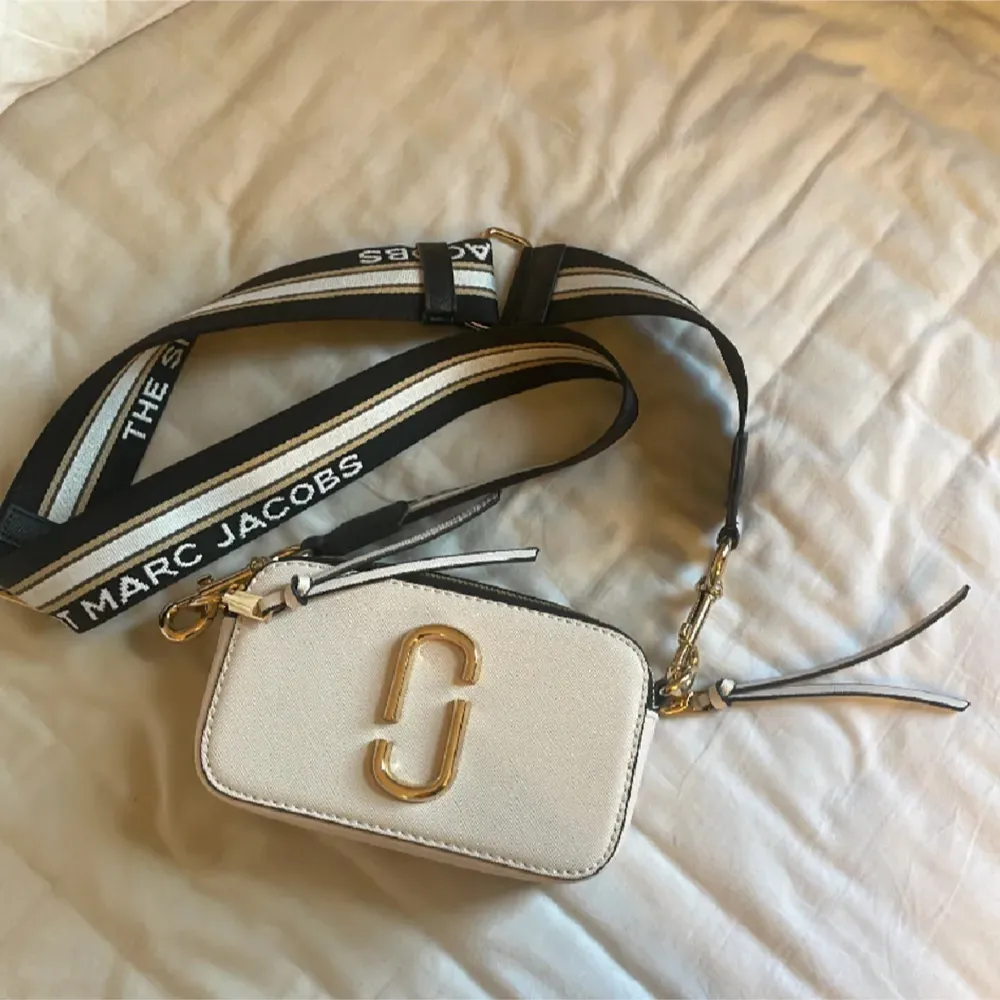 Snygg beige axelväska från Marc Jacobs med ett brett, justerbart axelband i svart och vitt. Väskan har en stilren design med guldfärgade detaljer och en praktisk dragkedja. Perfekt för att bära dina nödvändigheter med stil.. Laukut & Käsilaukut.