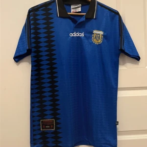 Argentina fotbollströja från Adidas - Snygg blå fotbollströja från Adidas med svart diamantmönster längs sidan och AFA-emblem på bröstet. Tröjan är kortärmad och tillverkad i 100% polyester, vilket gör den lätt och bekväm. Perfekt för fotbollsfans som vill visa sitt stöd.