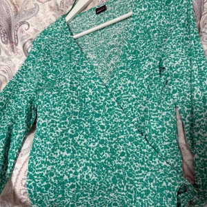 Grön mönstrad omlott topp - Snygg grön omlott topp från Gina Tricot med ett unikt mönster i vitt. Toppen har långa ärmar och en v-ringad design som ger en elegant look. Perfekt för att sticka ut med stil!
