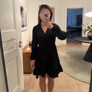Svart klänning med v-ringning - Snygg svart klänning med v-ringad hals och långa ärmar. Perfekt för en stilren look. Klänningen har en smickrande passform med resårband i midjan och faller fint i en kort längd.