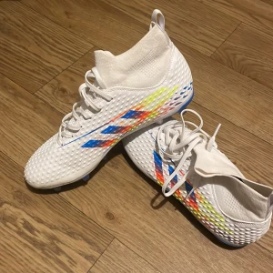 Vita fotbollsskor med färgglada detaljer - Snygga vita fotbollsskor med färgglada detaljer i blått, gult och rött. Skorna har snörning och en mönstrad yta för extra grepp. Perfekta för fotbollsplanen med sina blå dobbar.