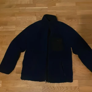 Mörkblå fleece jacka från Indicode jeans - Säljer en mörkblå fleece jacka från Indicode jeans med hög krage och dragkedja. Perfekt för kyligare dagar. Jackan har en stilren design.