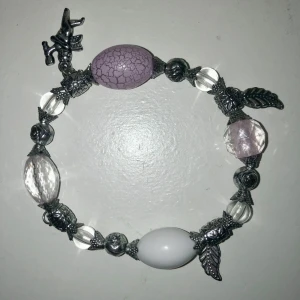Armband med charms och stenar - Charmigt armband med olika stenar i lila, vit och genomskinlig färg. Armbandet har detaljer som löv och rosor i metall. Perfekt för att ge en unik touch till din stil.