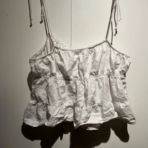 Vit ärmlös topp med knyt detalj framtill - Säljer en söt vit ärmlös topp med knyt detalj framtill och tunna axelband. Perfekt för sommardagar och kan enkelt matchas med jeans eller kjol. Axelbanden är justerbara för bästa passform.