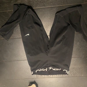 Svarta träningsleggings från Nike - Säljer ett par svarta träningsleggings från Nike med en vit logga på benet. De har en elastisk midja med texten 'Nike Pro'. Perfekta för träning och bekväma att bära.