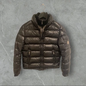 Brun dunjacka från Moncler - Snygg svart dunjacka från Moncler med hög krage och dragkedja framtill. Jackan har långa ärmar och en quiltad design som ger en stilren look. Perfekt för kyligare dagar. Storlek 152cm, den ser typ grön ut på bilden men den är svart