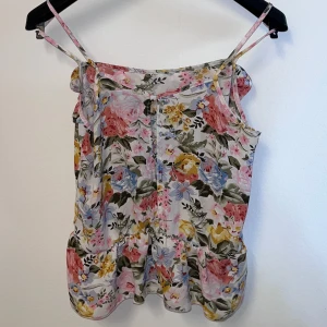 Blommig linnetopp från BikBok - Söt blommig linnetopp från BikBok med tunna axelband och volangdetaljer. Toppen har ett färgglatt mönster med rosa, blå och gula blommor på en vit bakgrund. Perfekt för varma sommardagar.