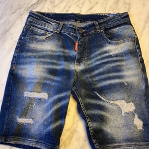 Blå jeansshorts från DSQUARED2 - Snygga blå jeansshorts från DSQUARED2 med slitna detaljer och en cool tvättad look. De har en knapp och dragkedja framtill samt klassiska fem fickor. Perfekta för en avslappnad stil.