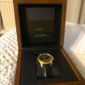 Elegant klocka från Hauger - Snygg klocka från Hauger med en stilren svart urtavla och guldfärgad boett. Klockan har ett svart läderarmband och levereras i en elegant trälåda. Perfekt för den som söker en tidlös accessoar.     Lägg bud!