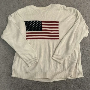 Vit tröja med amerikansk flagga från Focus America - Säljer en vit tröja från Focus America med en stor amerikansk flagga på ryggen. Tröjan har långa ärmar och är tillverkad i ett ribbat material. Perfekt för en avslappnad stil med en touch av patriotism.