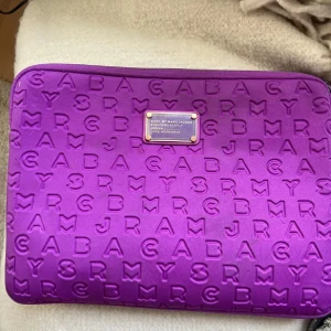 Lila datorväska från Marc by Marc Jacobs - Snygg lila datorväska från Marc by Marc Jacobs med präglad textdesign. Väskan har en dragkedja och är perfekt för att skydda din laptop med stil. Den är både praktisk och trendig, perfekt för dig som vill ha något unikt.