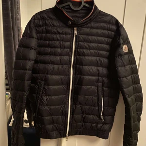 Svart dunjacka från Moncler - Säljer en stilren svart dunjacka från Moncler med dragkedja och ribbade detaljer. Jackan har en quiltad design och långärmade ärmar. Perfekt för kyligare dagar.