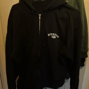 Svart zip up hoodie med tryck sportig - Snygg svart hoodie med dragkedja och tryck på både fram- och baksidan. Texten 'Speedway Champ' pryder framsidan medan baksidan har ett större tryck med racingtema. Perfekt för en avslappnad stil.