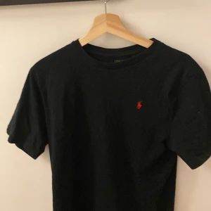 Svart t-shirt från Ralph Lauren - Snygg svart t-shirt från Ralph Lauren med det klassiska röda broderade logotypen på bröstet. Perfekt för en stilren och avslappnad look. Tillverkad i mjuk bomull för extra komfort. Storlek S/M