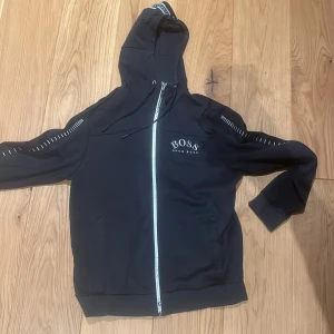 Mörkblå huvtröja från Boss - Säljer en stilren svart huvtröja från Boss med dragkedja och logotyp på bröstet. Tröjan har långa ärmar med vita detaljer längs ärmarna och en justerbar huva. Perfekt för en casual look.