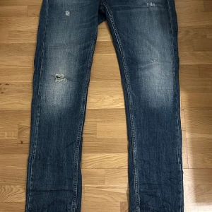 Blå jeans från Replay - Säljer ett par blå Replay jeans i modellen Grover. De har en snygg slitning och en klassisk femficksdesign med knappar och dragkedja. Perfekta för en avslappnad stil.