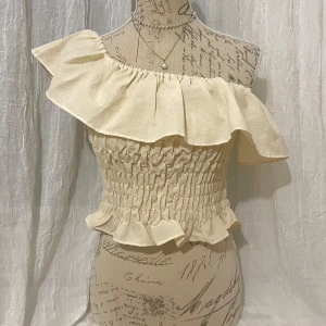 Beige offshoulder topp med volang - Säljer en söt beige offshoulder topp med volangdetaljer. Man kan ha den på sig på olika sätt. Den är stretchig så passar bra S-M. Dock är det lite lös vid midjan (se sista bild) så om du har smal midja (under 65cm) kan det tyvärr inte sitta perfekt på dig.