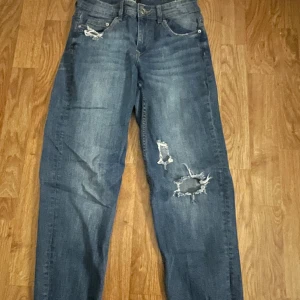 Blå jeans med slitningar - Säljer ett par blå jeans med coola slitningar på framsidan. De har en klassisk femficksdesign och en bekväm passform. Perfekta för en avslappnad stil.