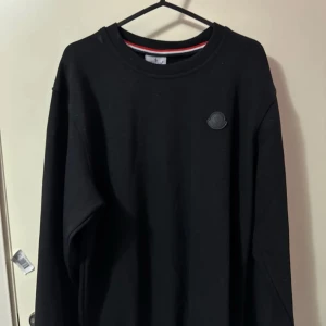 Moncler tröja  - Säljer en stilren svart långärmad tröja från Moncler med rund halsringning. Tröjan har en diskret Moncler-logga på bröstet och en röd-vit randig detalj vid halsen. Perfekt för en casual look.