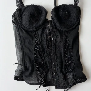 Svart korsett med spets och snörning - Snygg svart korsett med spetsdetaljer och snörning fram och bak. Den har justerbara axelband och hakknäppning framtill. 