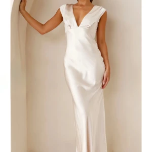 Champagnefärgad långklänning - Elegant champagnefärgad långklänning i satin med djup v-ringning och öppen rygg. Klänningen har en figurnära passform och korta ärmar. Perfekt för festliga tillfällen.