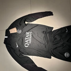 Svart långärmad PSG fotbollströja och byxor - Snygg svart långärmad PSG fotbollströja med matchande byxor från Nike. Tröjan har en dragkedja vid halsen och PSG-loggan på bröstet. Byxorna har elastisk midja och PSG-loggan på benet. Perfekt för träning eller avslappnad stil.