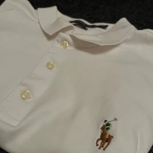 Vit pikétröja från Ralph Lauren - Säljer en klassisk vit pikétröja från Ralph Lauren i slim fit. Tröjan har korta ärmar och tre knappar framtill. Den är dekorerad med det ikoniska broderade polospelar-emblemet på bröstet. Perfekt för en stilren look! Passar även S 
