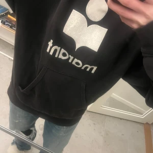 Svart hoodie från Isabel Marant - Säljer en svart hoodie från Isabel Marant med en stor vit logotyp på framsidan. Hoodien har en rymlig passform och en praktisk magficka. Perfekt för en avslappnad stil.