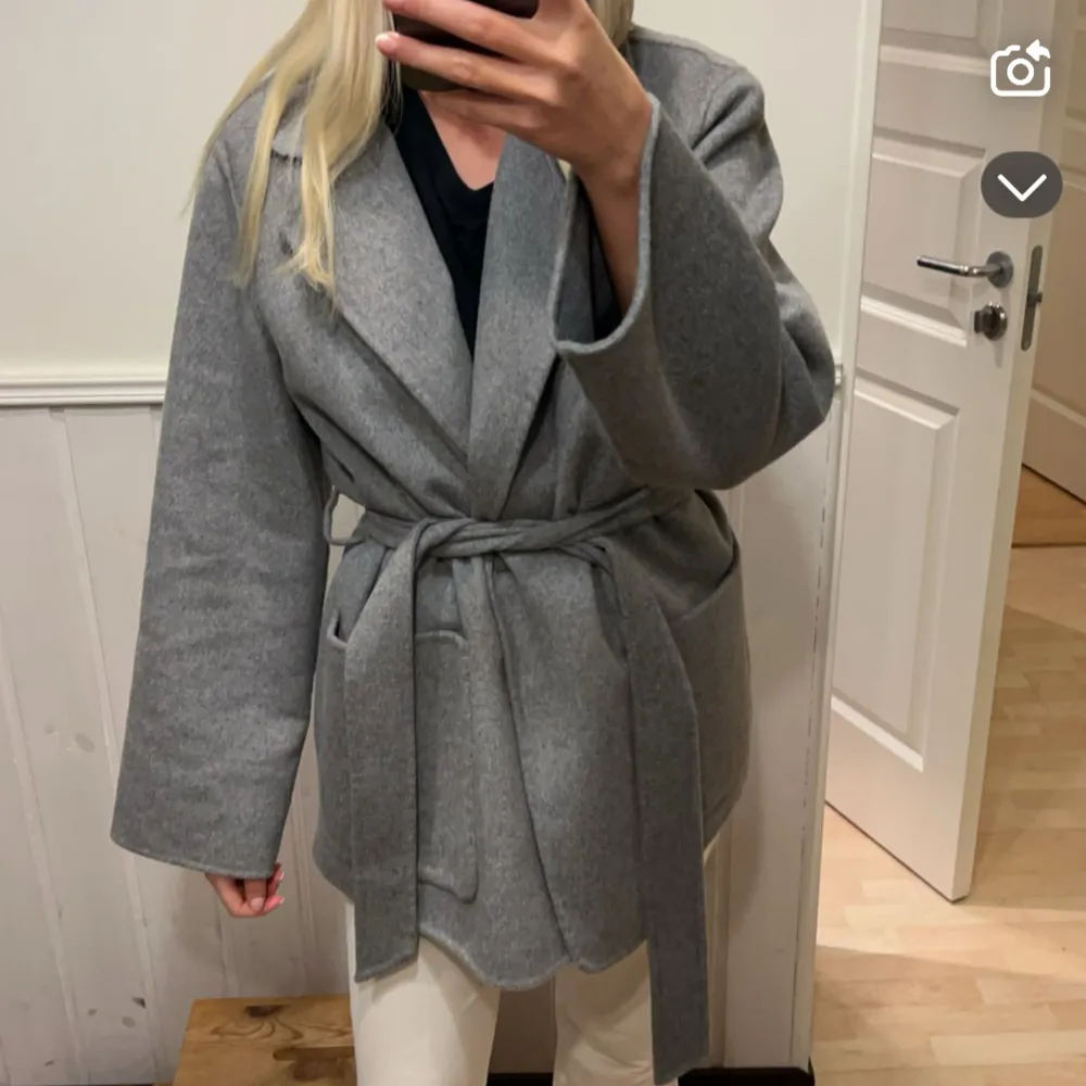 Säljer en stilren grå kappa från Stockh LM. Den har en klassisk design med knytskärp i midjan och stora fickor framtill. Perfekt för kyligare dagar med sin långa ärm och mjuka ullblandning. En tidlös favorit i garderoben!. Takit.