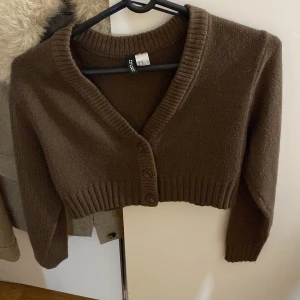 Brun croppad kofta från H&M Divided - Säljer en brun croppad kofta från H&M Divided. Koftan har en djup v-ringning och knappar framtill. 