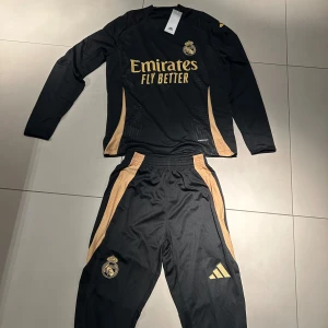 Svart långärmad fotbollströja från Adidas - Snygg svart långärmad fotbollströja från Adidas med guldiga detaljer och Real Madrid-logga. Perfekt för fotbollsfans som vill ha stil på planen. Tröjan har en modern design med 'Emirates Fly Better' tryckt på framsidan.