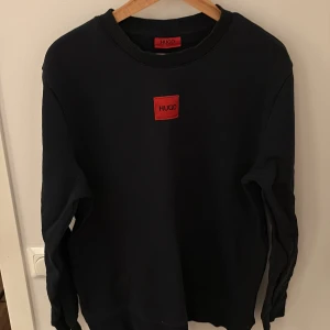 Svart tröja från Hugo Boss - Snygg svart tröja från Hugo Boss med röd logga på bröstet. Tröjan har en klassisk rund hals och långa ärmar, perfekt för en stilren look. Passar bra till både jeans och chinos.