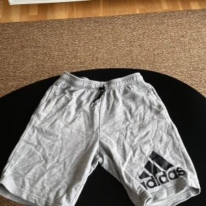 Grå shorts från Adidas - Säljer ett par grå shorts från Adidas med elastisk midja och dragsko. De har en stor svart Adidas-logga på ena benet. Perfekta för en avslappnad stil eller träning.