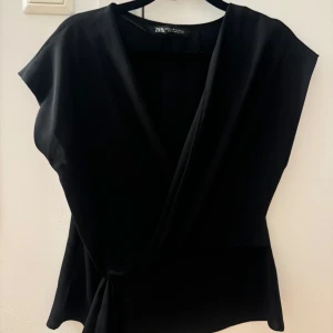 Svart omlottopp från Zara - Snygg svart omlottopp från Zara med korta ärmar och knytning i sidan. Perfekt för en stilren look.