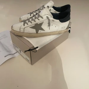 Golden Goose Superstar - Snygga vita sneakers från Golden Goose med en grå stjärna på sidan och mörkblå häl. Skorna har snörning och en klassisk design, har slitningar som är meningen. Allt kommer med som box, dustbag, alla tags, kvitto digitalt och i papperform. Är i jättebra skick! 9/10,  enda defekten är att de är hål i hälen men man märker inte av det när man har på skorna överhuvudtaget. Skriv vid funderingar!